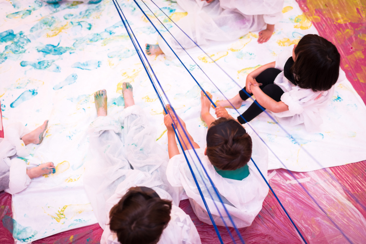 Vista posterior de varias niñas con plásticos protectores sentadas sobre una tela con pintura.